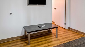 Una habitación con piso de madera, una mesita con un control remoto, un televisor de pantalla plana montado en la pared y una puerta con interruptor de luz.