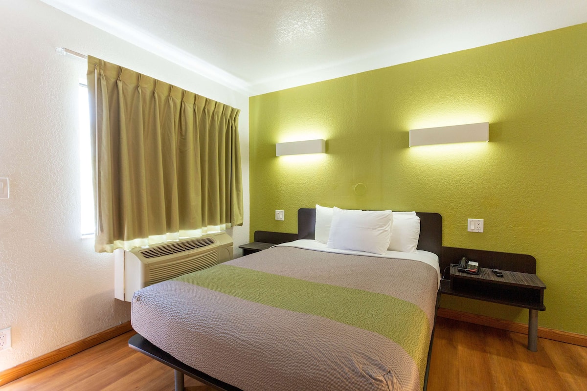 A hotel room with a bed, bedside tables, a window with curtains, and a wall-mounted light fixture.