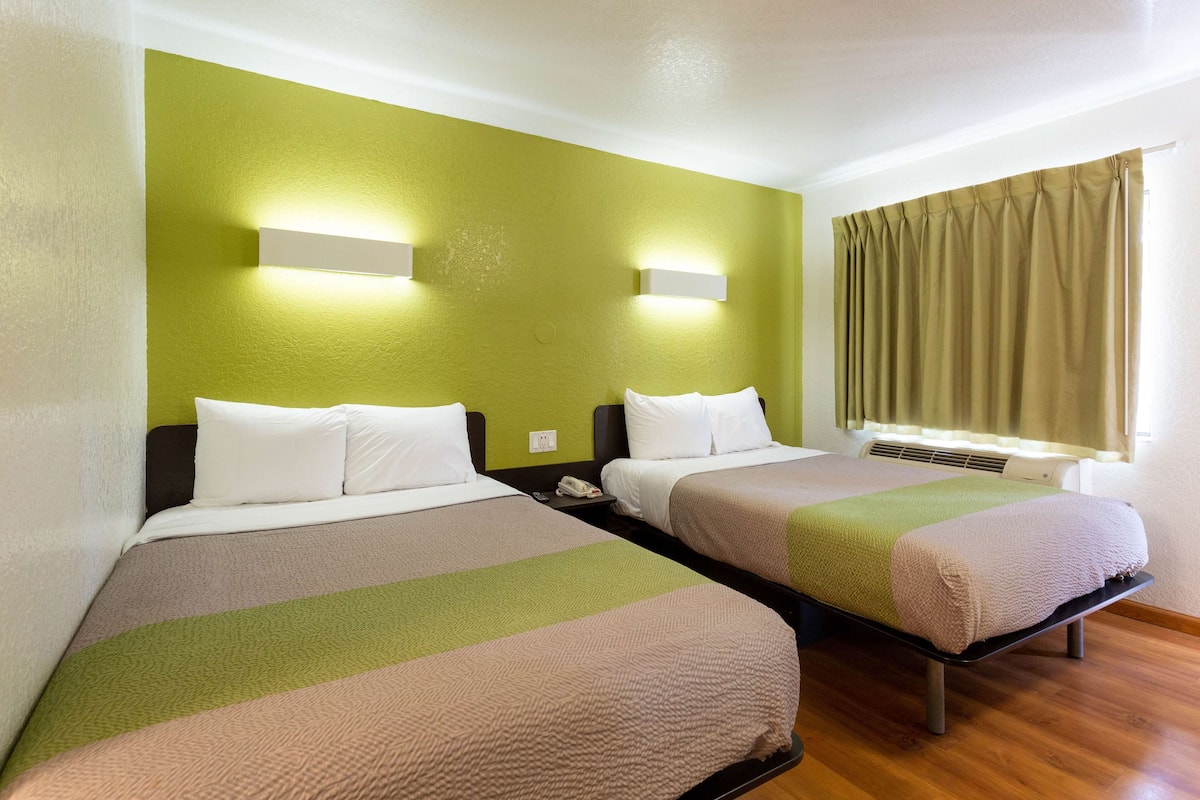 A hotel room with two beds, each with a pillow and a blanket. There is a nightstand with a phone and a wall-mounted light.