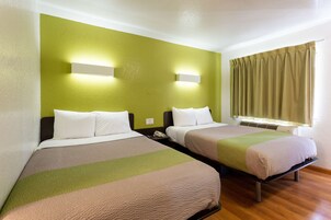 Habitación de hotel con dos camas, cada una con una almohada y una manta. Hay una mesita de noche con un teléfono y una lámpara fijada a la pared.