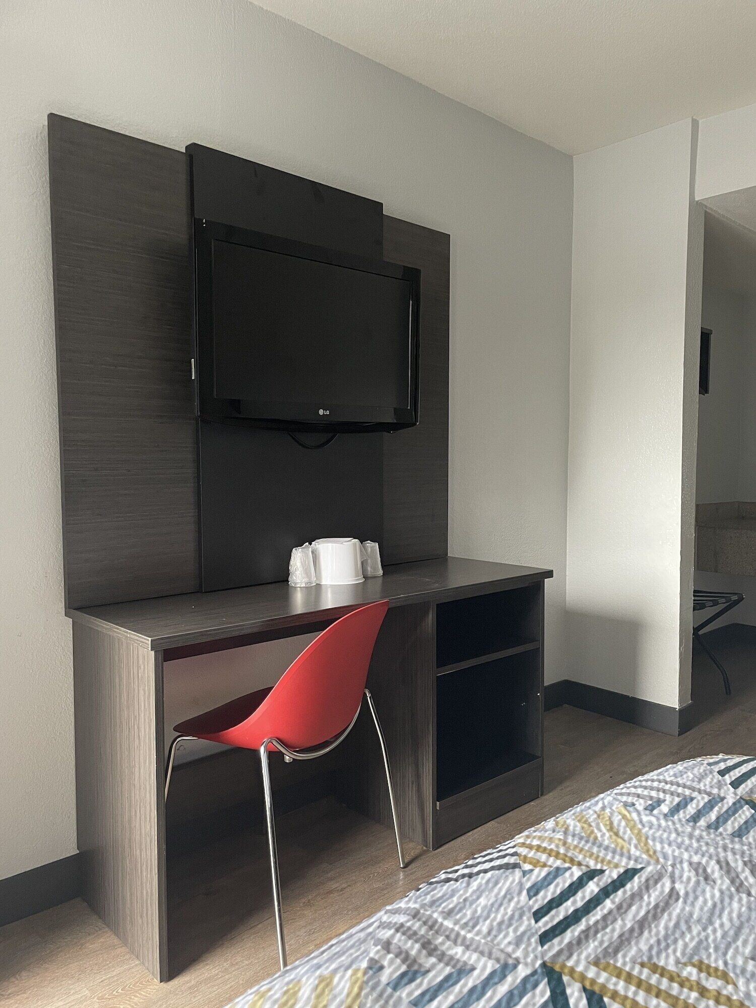 A hotel room with a dark wood desk, a red chair, a flat-screen TV mounted above the desk, and a bed with a patterned bedspread.