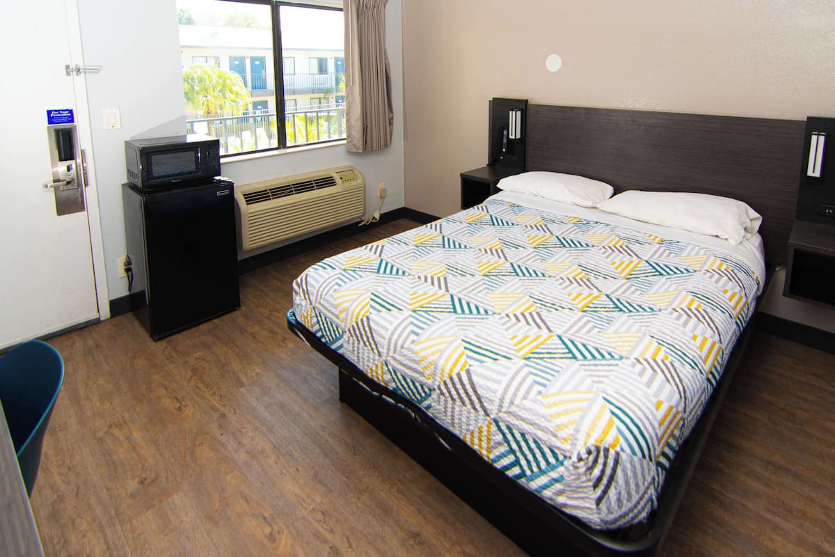 A hotel room with a bed, a desk with a microwave, a heater, and a window with a view of buildings.