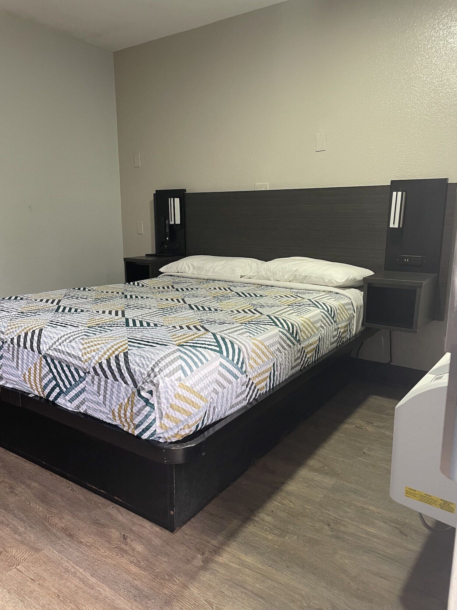 Uma cama com colcha estampada, cabeceira escura e mesa de cabeceira com abajur.