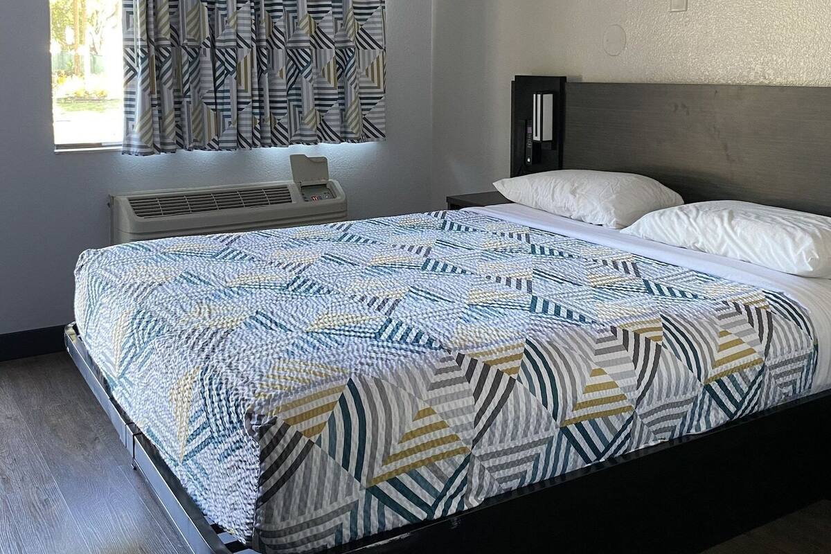 A bedroom with a bed, a window with patterned curtains, a bedside table, and a wall-mounted heater.