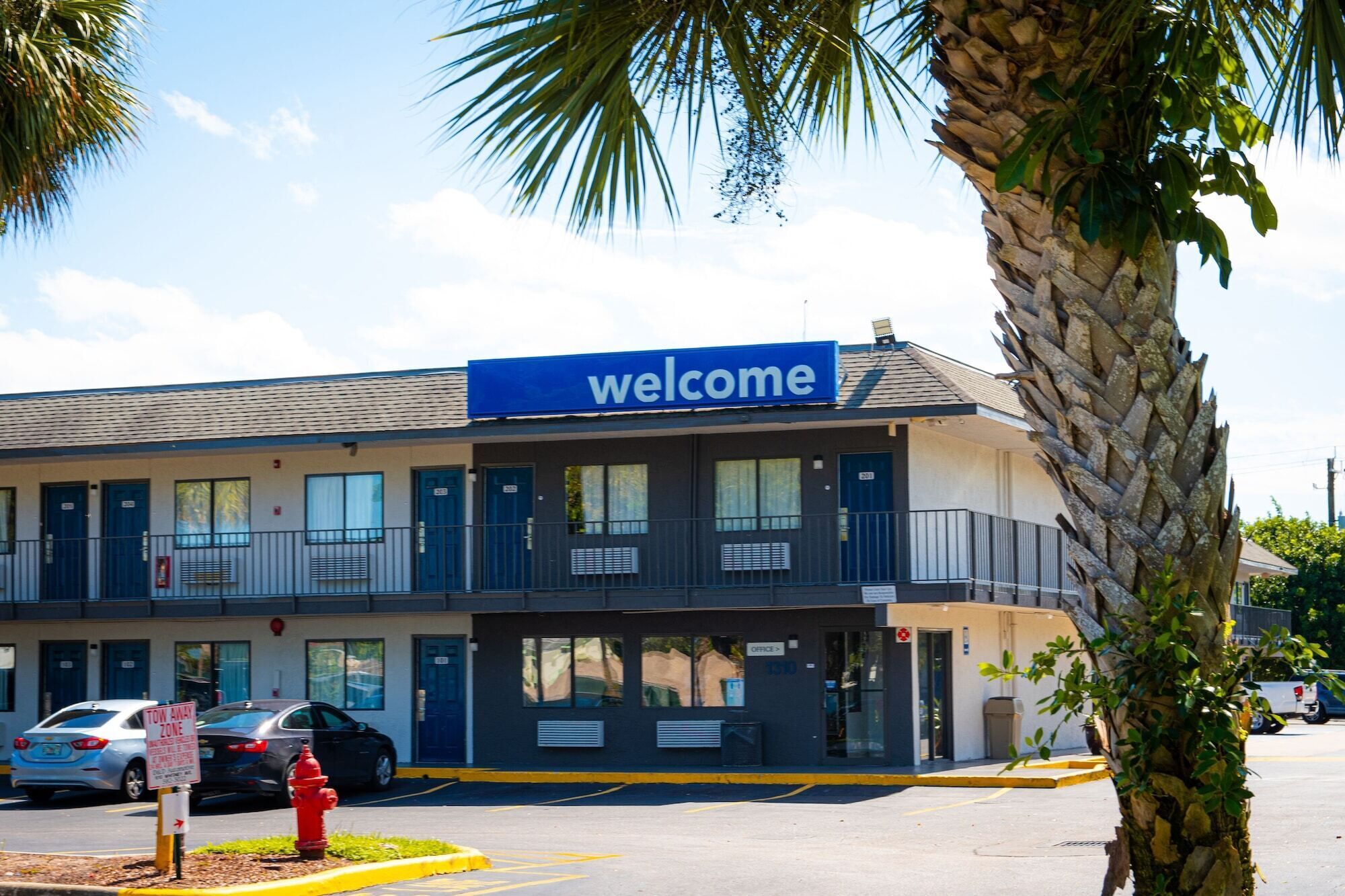 A two-story building with a 'Welcome' sign, a parking lot, and a red fire hydrant.