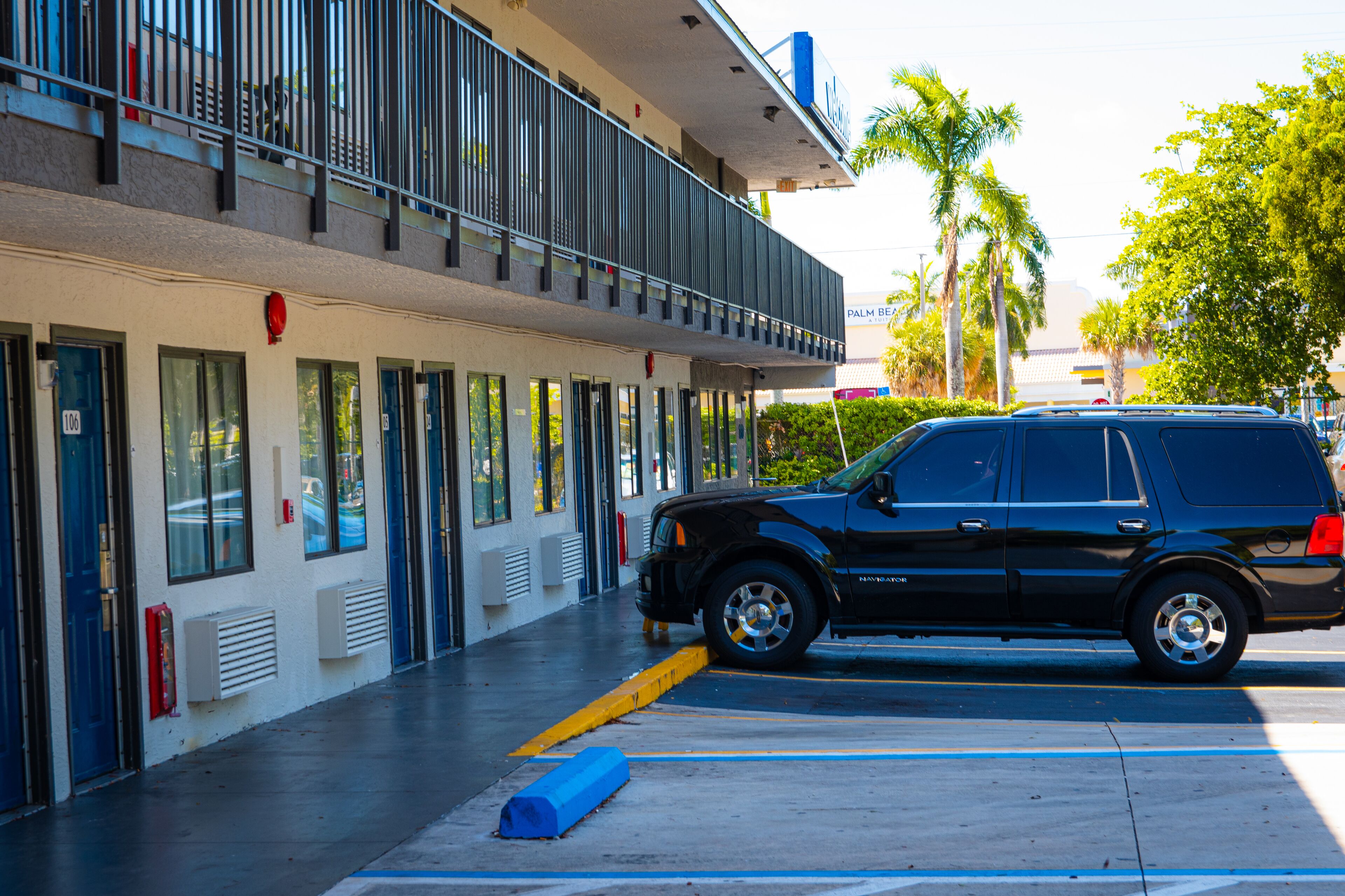 A black SUV parked in front of a building with multiple doors and windows, a balcony, and a palm tree in the background.