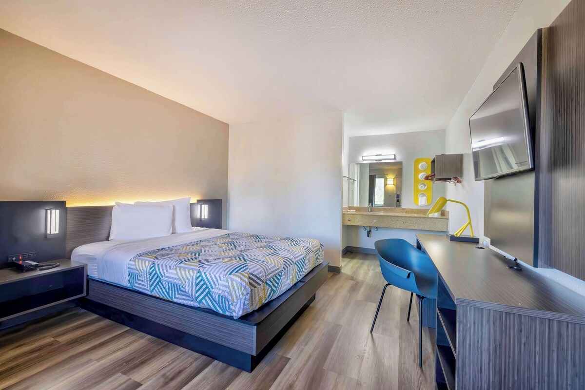 A modern hotel room with a bed, bedside table, desk, chair, and a TV.