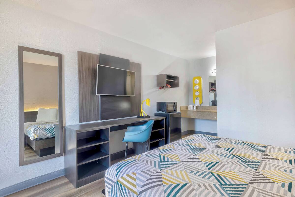 A compact hotel room with a bed, a desk with a chair, a TV, and a small kitchenette.