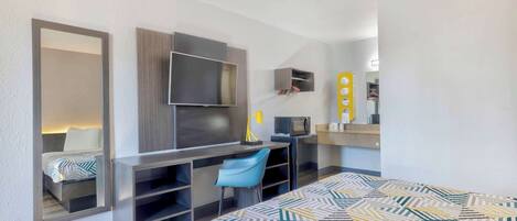 Una habitación de hotel compacta con cama, escritorio, silla, televisión y una pequeña kitchenette.