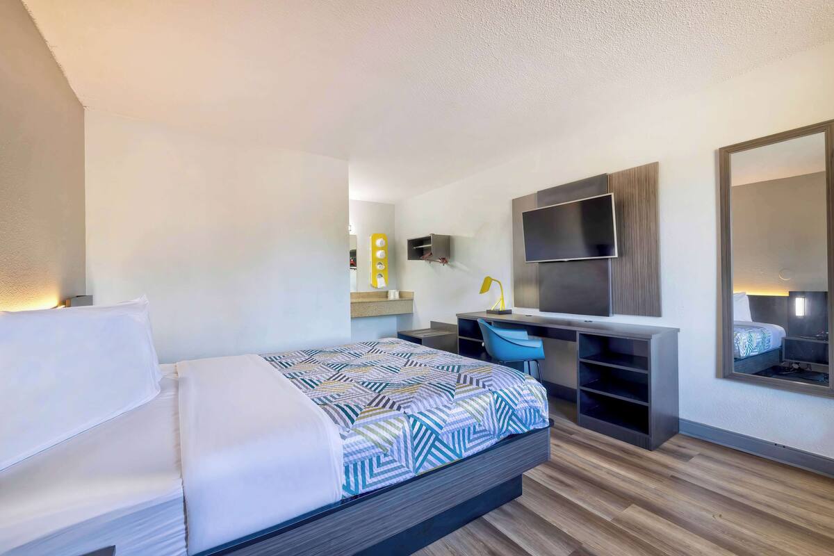 A modern hotel room with a bed, desk, chair, TV, and a large mirror.