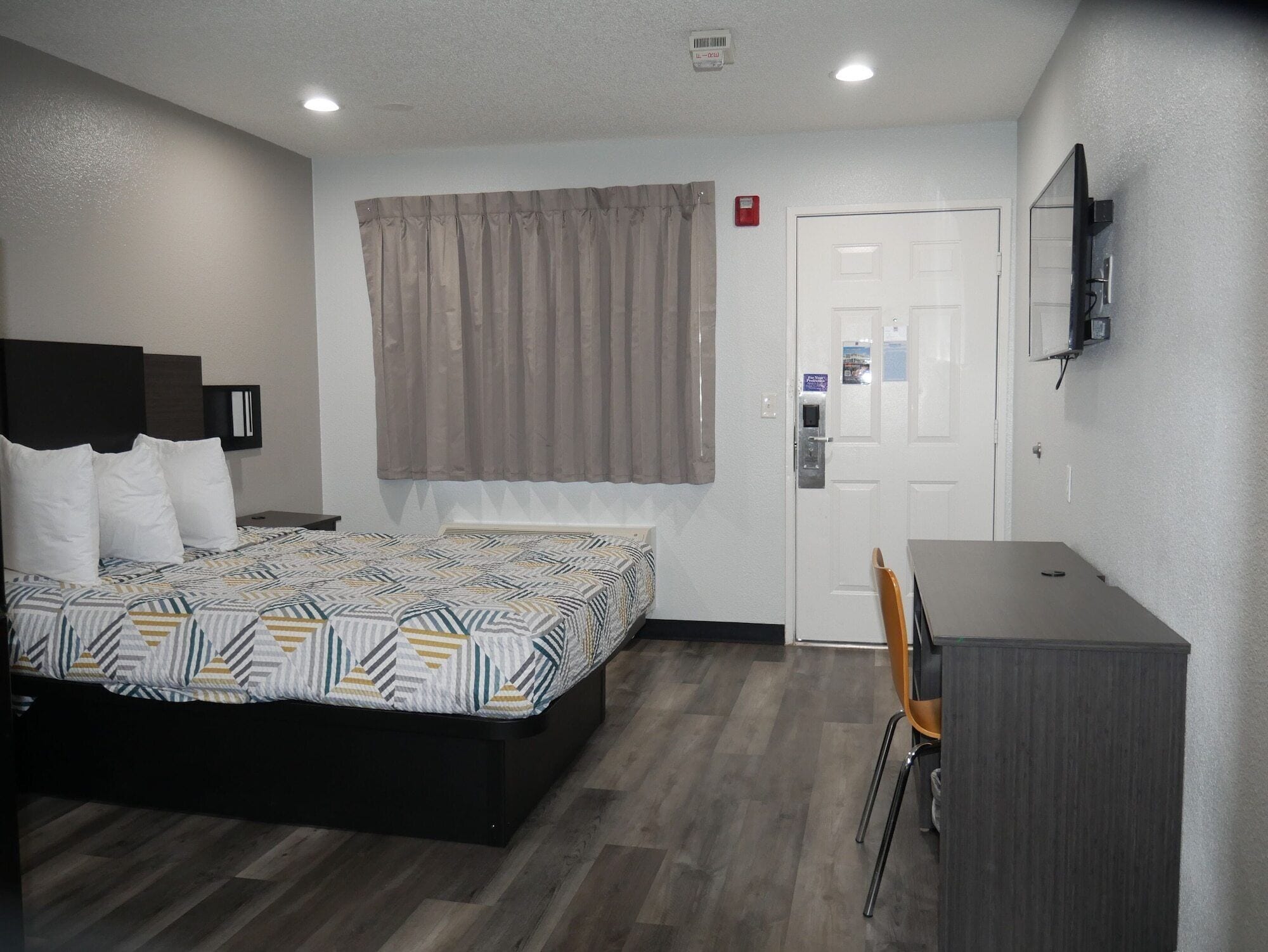 Habitación de hotel con cama, escritorio, silla, televisión y puerta con panel de seguridad.