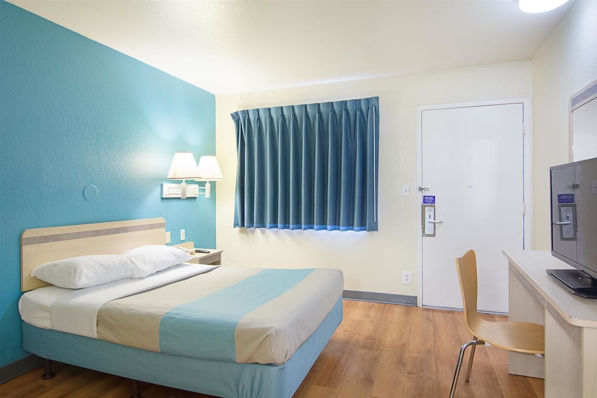 Hotellihuone, jossa on sänky, työpöytä kannettavalla tietokoneella, tuoli, ikkuna verhoilla, seinään kiinnitetty lamppu ja lukittava ovi.