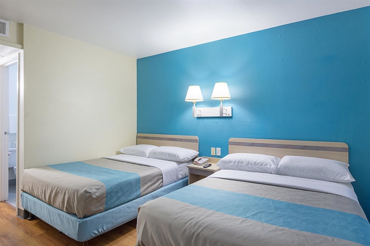 Hotellihuone, jossa on kaksi sänkyä, sininen tehosteseinä ja yöpöytä, jossa on lamppu.