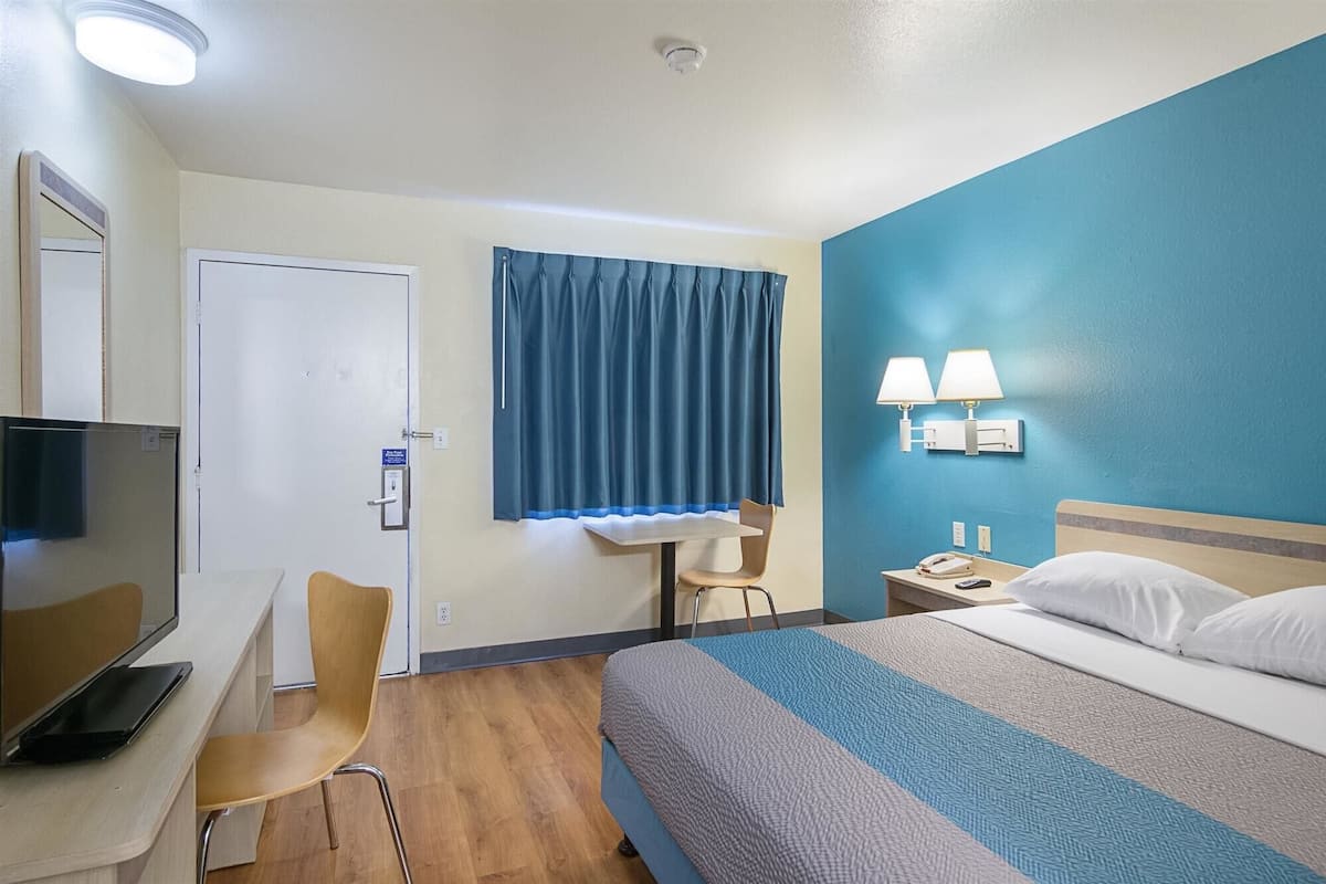 Hotellihuone, jossa on sänky, työpöytä tuolilla, televisio ja ikkuna, jossa on verhot.