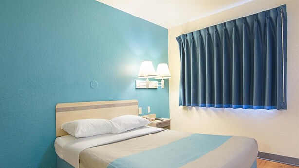 Una habitación de hotel con una cama, una mesita de noche, una lámpara y una ventana con cortinas.
