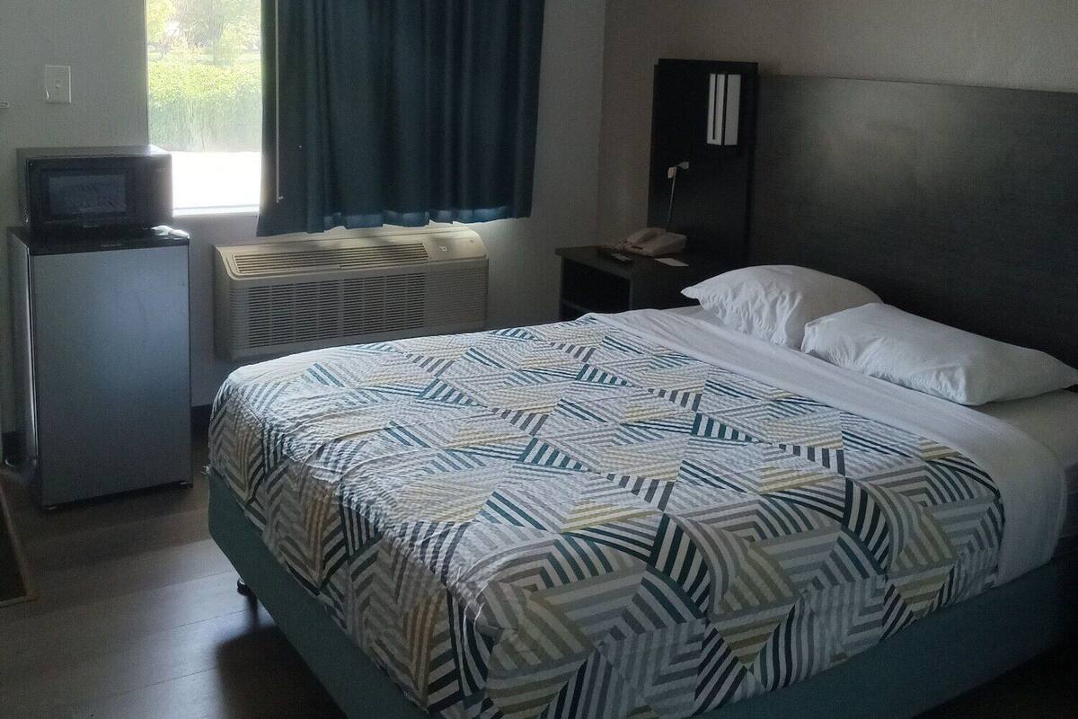 Una habitación de hotel con cama, escritorio con computadora, ventana con cortinas y una lámpara en el techo.