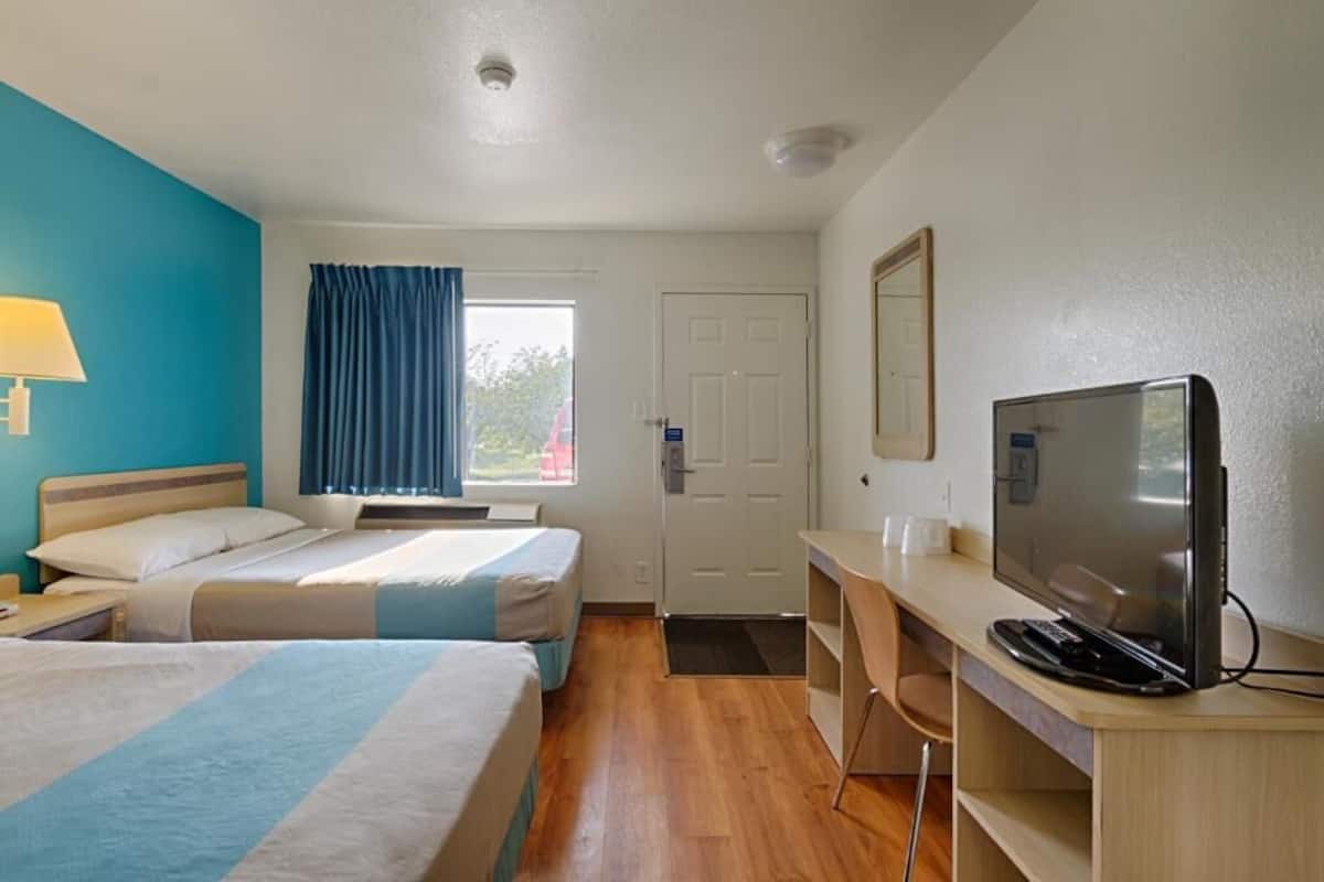 Habitación de hotel con dos camas, un escritorio con televisor, una ventana con cortinas y una puerta.