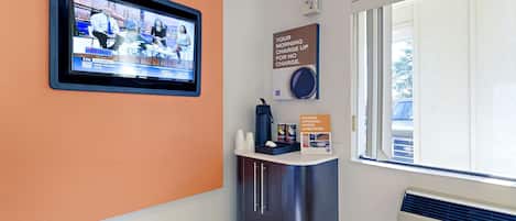 A hotel reception area with a television displaying a news program, a coffee machine, and a sign about morning charges.