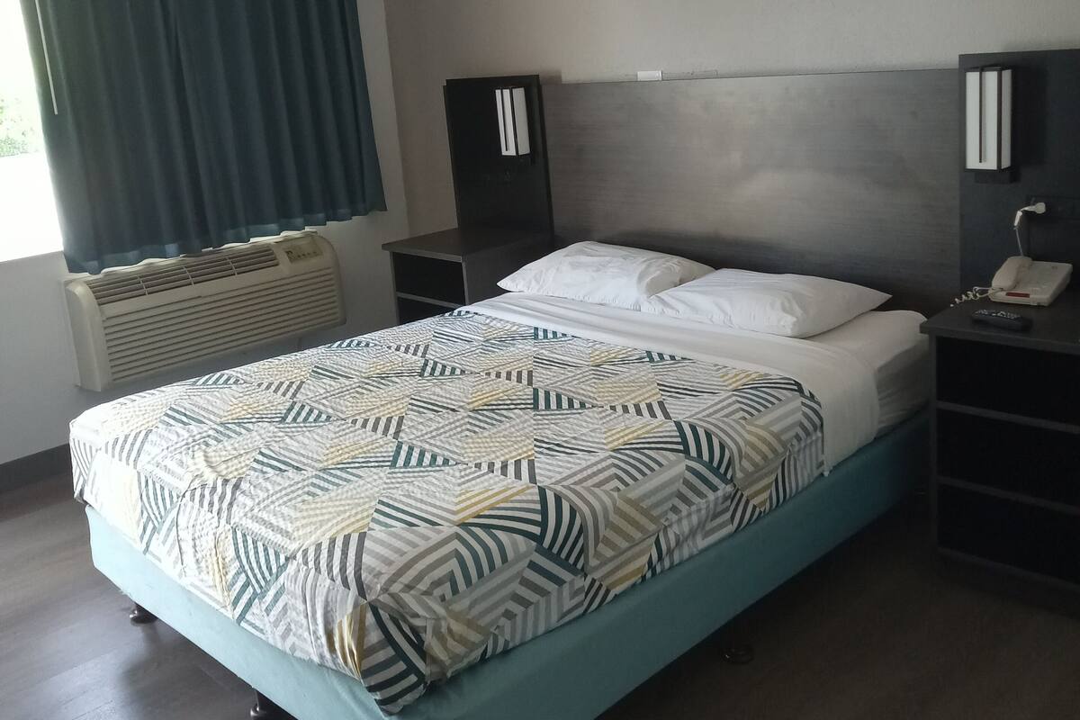 Una habitación de hotel moderna con una cama, una mesita de noche con teléfono, una lámpara de pared y un ventanal con cortinas.