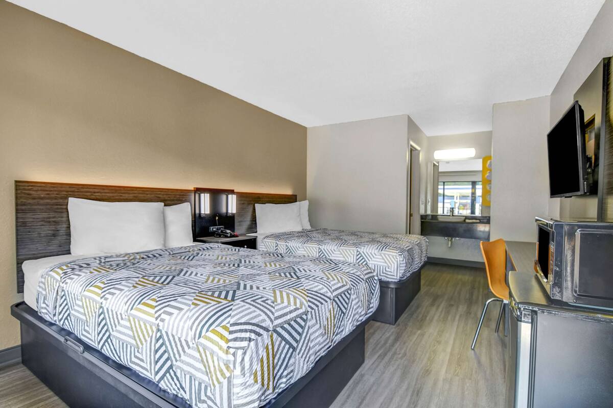 A hotel room with two beds, a desk, a chair, a TV, and a bathroom visible in the background.