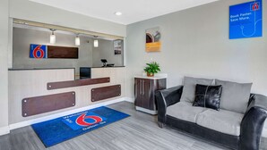 A modern reception area with a desk, a couch, a plant, and a welcome mat. - Motel 6 Dalton, GA (Dalton)