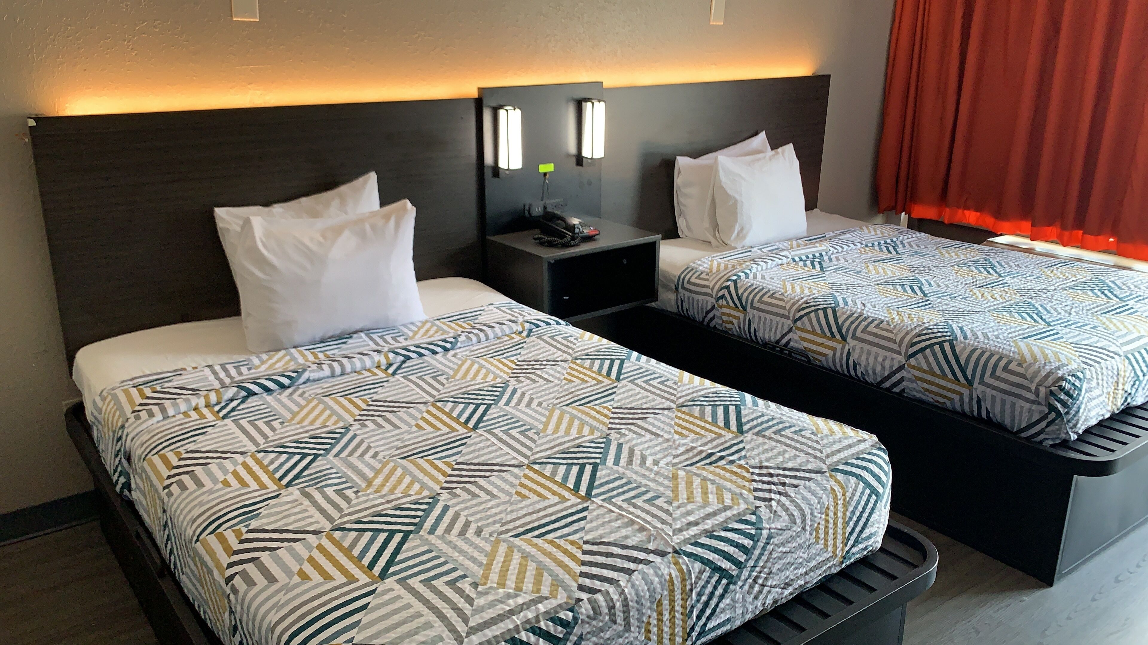 A hotel room with two beds, each with patterned bedding and white pillows. There is a nightstand with a lamp and a phone between the beds.