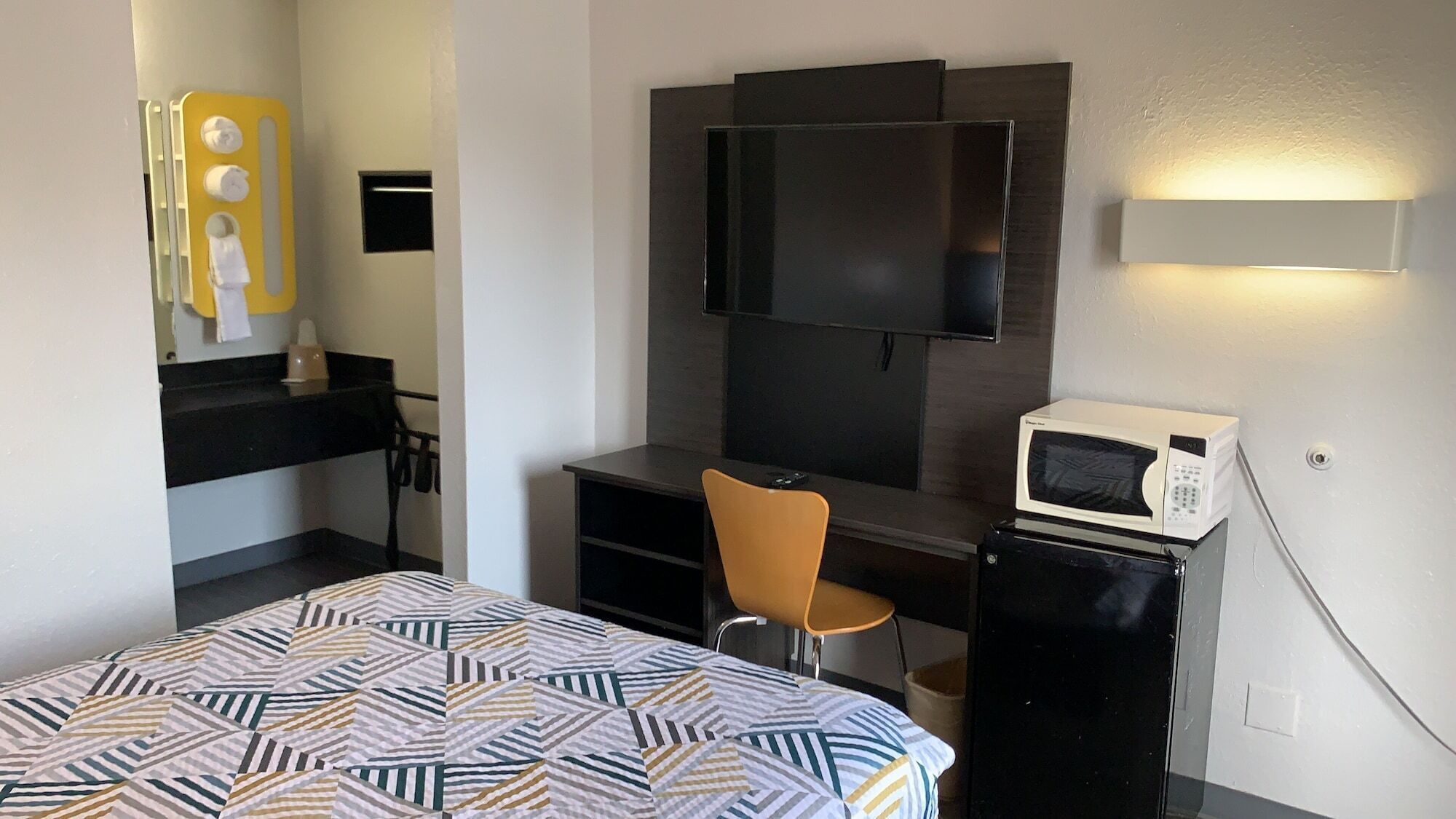 A hotel room with a bed, a desk with a chair, a television, a microwave, and a small shelf with toiletries.