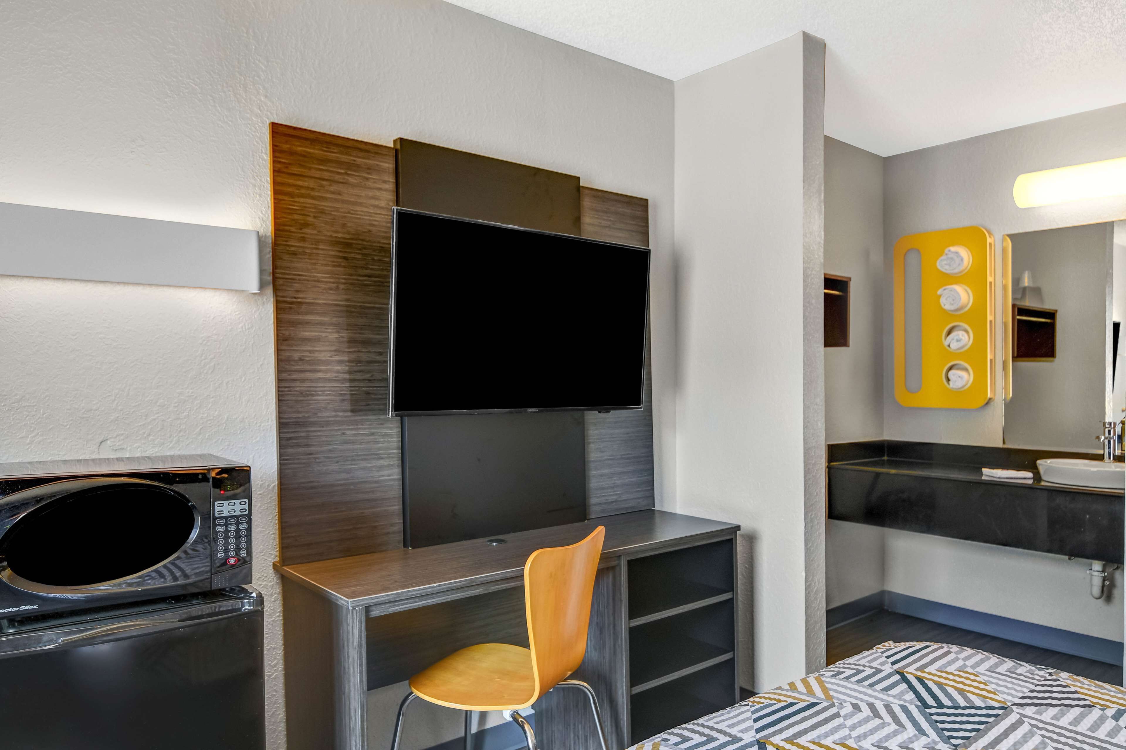 Habitación de hotel con televisión de pantalla plana montada en la pared, microondas, una silla de madera y baño con lavamanos y espejo.