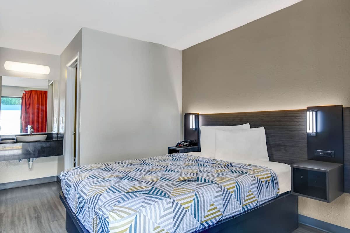 A hotel room with a large bed, a bedside table with a lamp, a modern bathroom with a sink and mirror, and a red curtain.