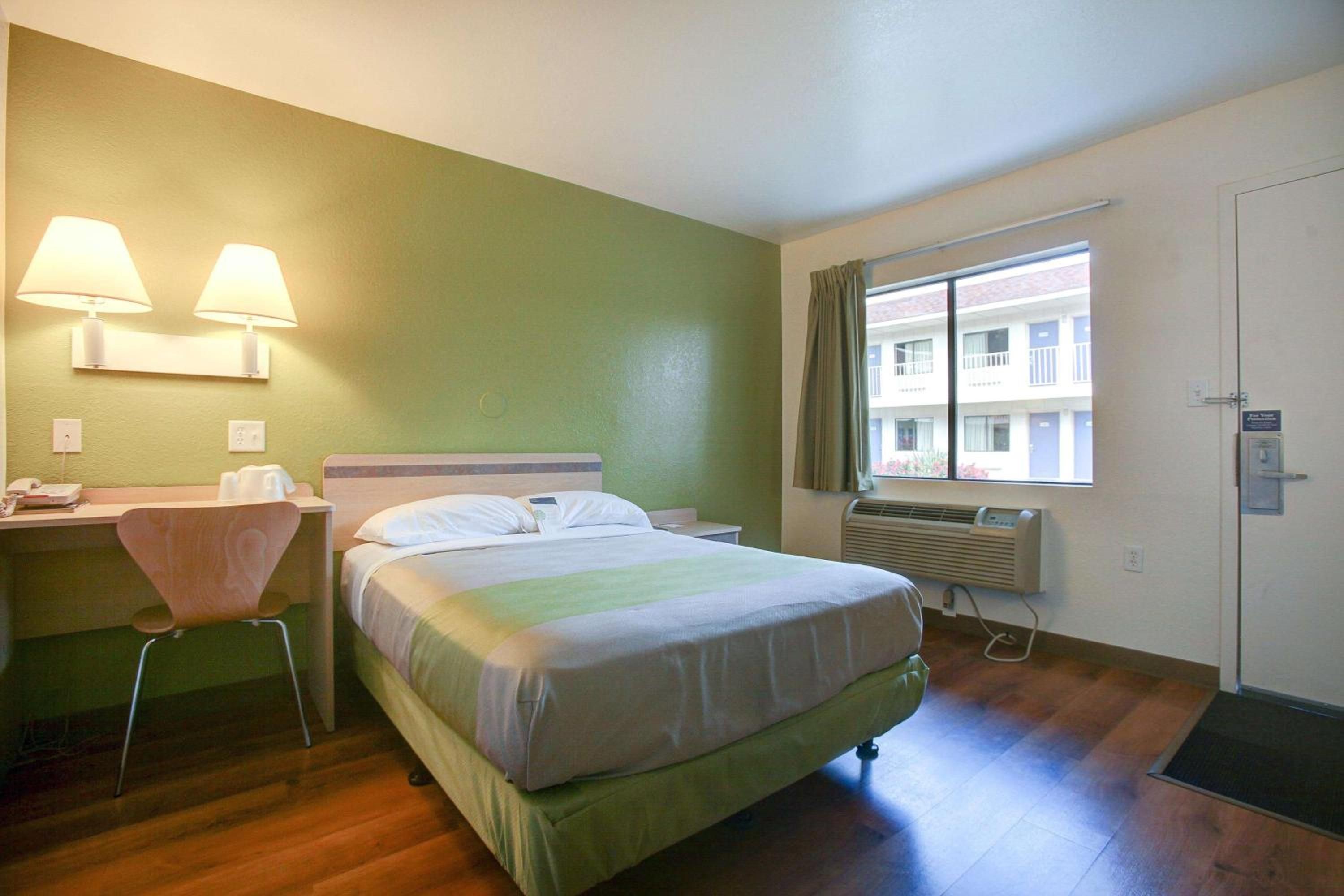 Quarto de hotel com cama, escrivaninha com cadeira, duas luminárias de parede, janela com cortinas e aparelho de ar condicionado.