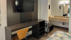 A hotel room with a flat-screen TV mounted on a dark wood panel, a desk with a chair, a large mirror above a sink, and a bed with patterned bedding. - Motel 6 Elkton, MD (Elkton)