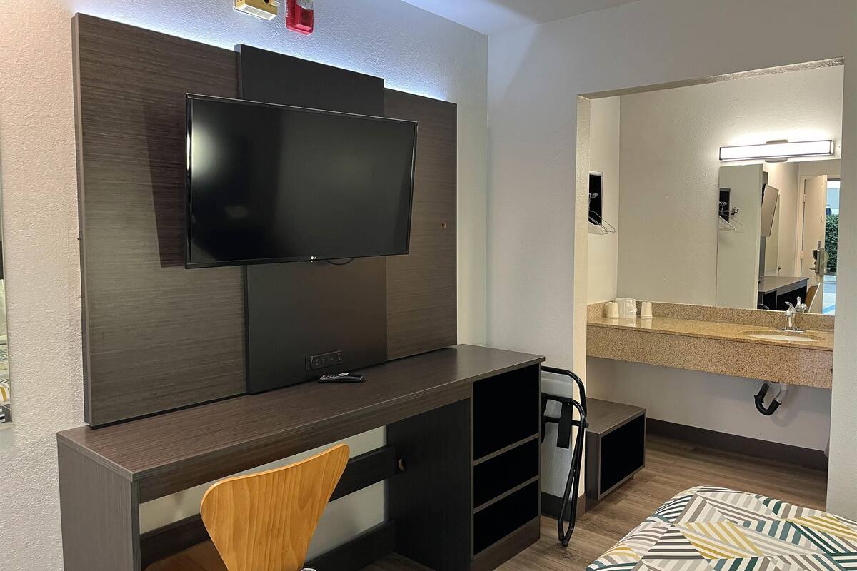 Ein Hotelzimmer mit Flachbildfernseher an einer dunklen Holzverkleidung, einem Schreibtisch mit Stuhl, einem großen Spiegel über einem Waschbecken und einem Bett mit gemusterten Betttextilien.