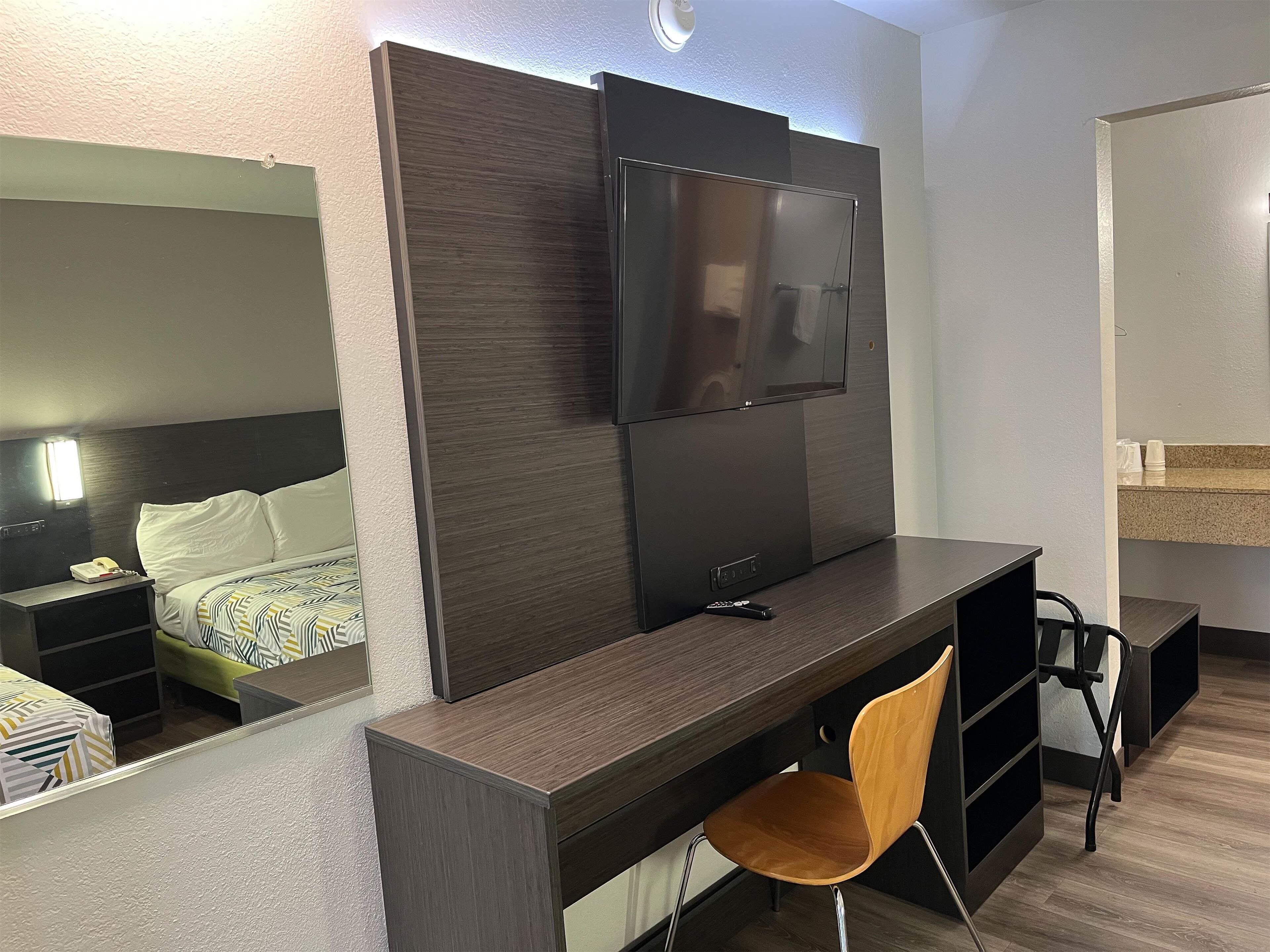 Une chambre d’hôtel avec un bureau en bois foncé, un téléviseur fixé au mur, une chaise, un lit avec une literie à motifs et une petite table de chevet avec une lampe.
