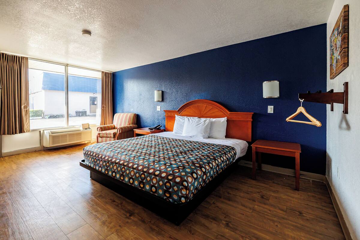 Une chambre d’hôtel avec un grand lit, une table de chevet en bois, une chaise, une fenêtre avec des rideaux et un luminaire fixé au mur.