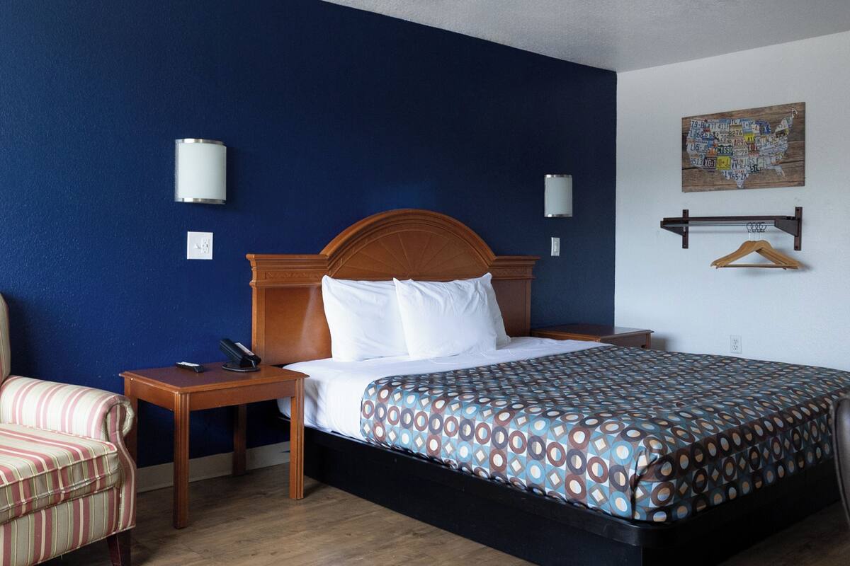 Une chambre d’hôtel avec un lit doté d’une tête de lit en bois, une literie blanche, une couvre-lit à motifs, une table de chevet en bois, un fauteuil à rayures et une carte accrochée au mur.