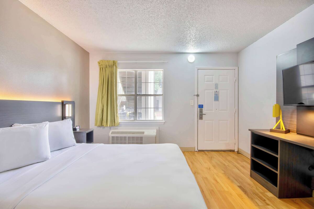 A hotel room with a bed, a window with curtains, a door, a TV, and a desk with a lamp.