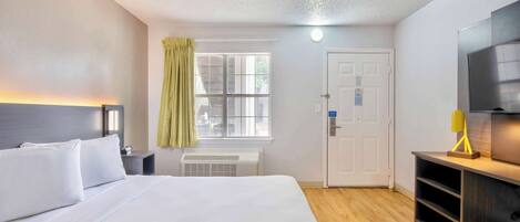 A hotel room with a bed, a window with curtains, a door, a TV, and a desk with a lamp.