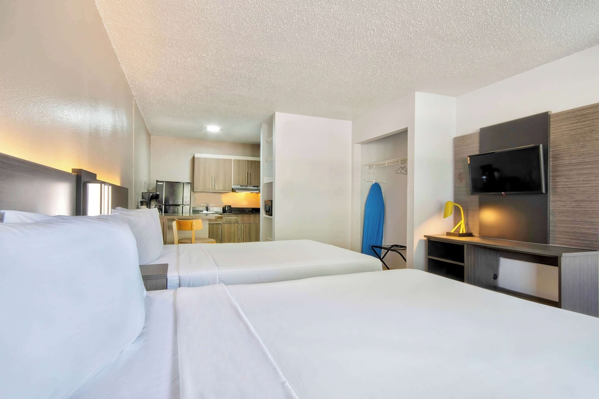 A hotel room with two beds, a TV, a desk, and a kitchenette in the background.
