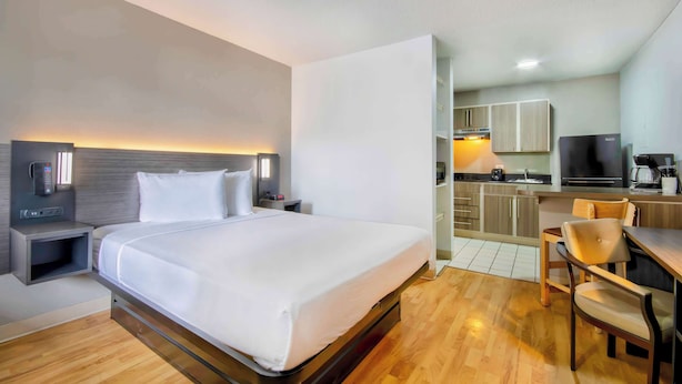 Een hotelkamer met een bed, nachtkastje en een klein bureau met stoel. Er is een kitchenette met een koelkast, magnetron en fornuis.
