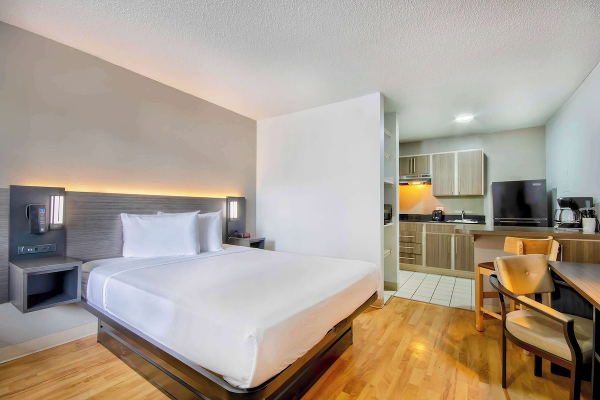 A hotel room with a bed, bedside table, and a small desk with a chair. There is a kitchenette with a refrigerator, microwave, and stove.