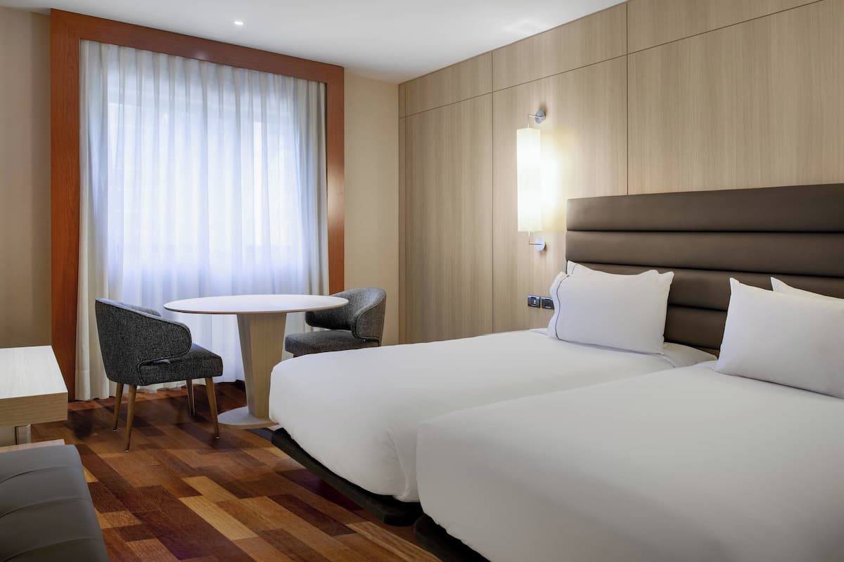 A hotel room with two beds, a small round table, a chair, and a large window with sheer curtains.