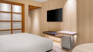 Una habitación de hotel moderna con una cama grande, un escritorio pequeño con una silla, un televisor montado en la pared y una puerta corredera con paneles de vidrio esmerilado.