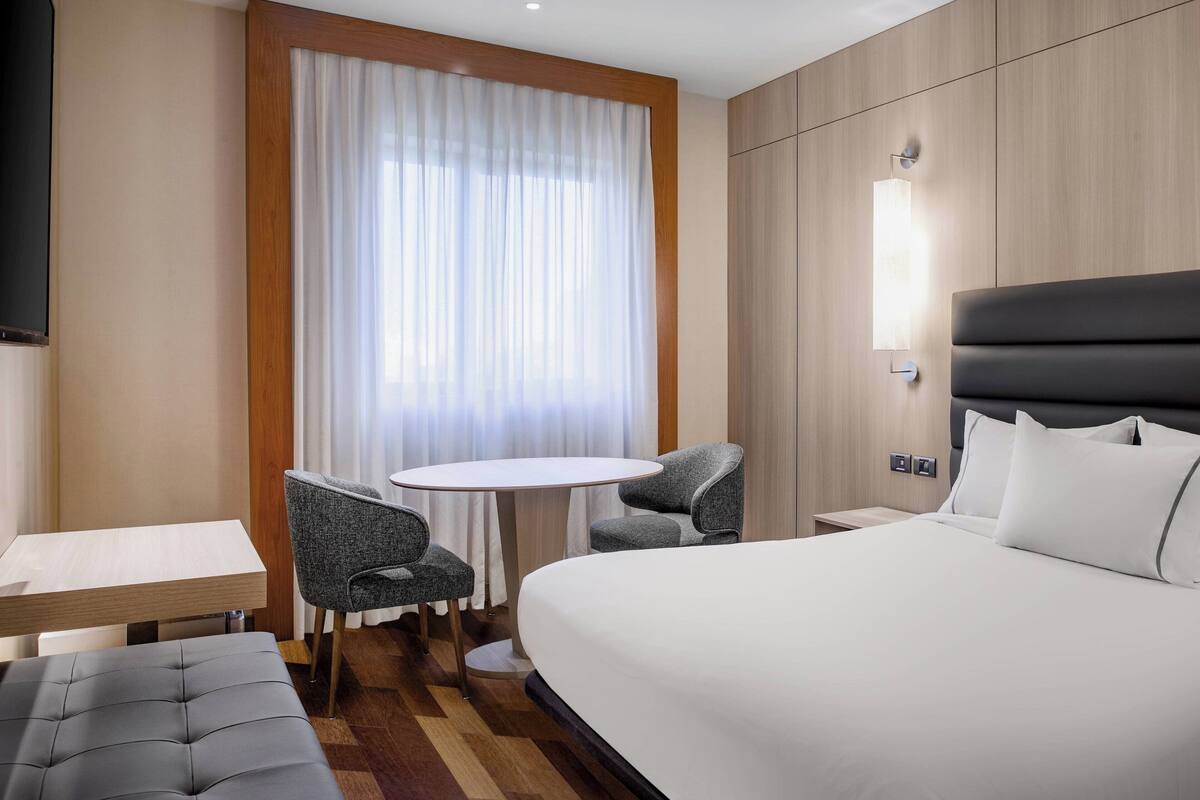 A hotel room with a large bed, a small round table, two chairs, a TV, and a window with curtains.