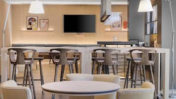 A modern hotel lobby with a central bar area, multiple seating options, and a large flat-screen TV mounted on the wall.