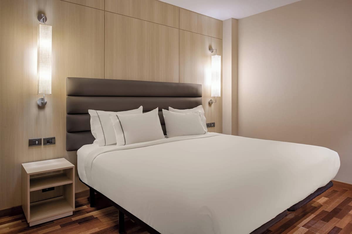 A modern hotel room with a large bed, a nightstand, and wall-mounted lighting.