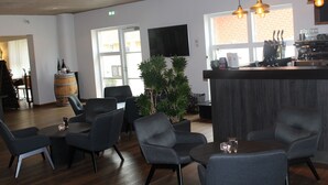 A modern bar area with a wooden ceiling, a large window, and a bar counter with a coffee machine. There are several chairs and a potted plant in the seating area. - Hotel Hedemarken (Grindsted)