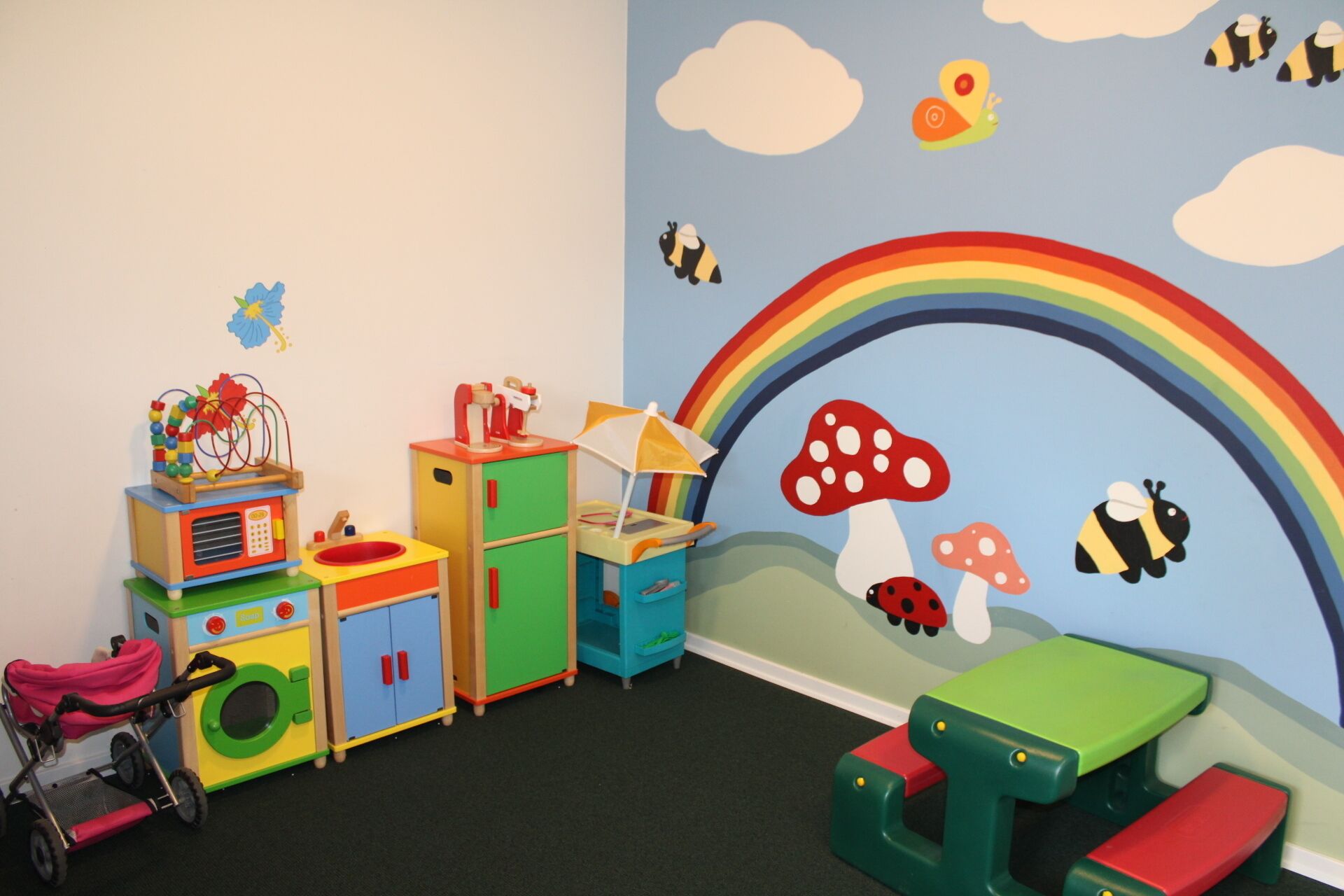A children's playroom with colorful toys, a toy kitchen, and a mural with a rainbow and insects.