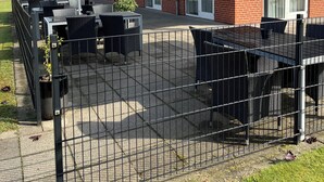 En udendørs patio med borde og stole, et hegnede område og en bygning med murstensfacade og store vinduer.