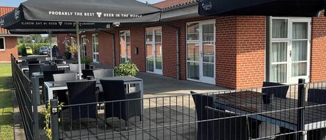 An outdoor patio area with tables and chairs, a fenced enclosure, and a building with a brick facade and large windows.