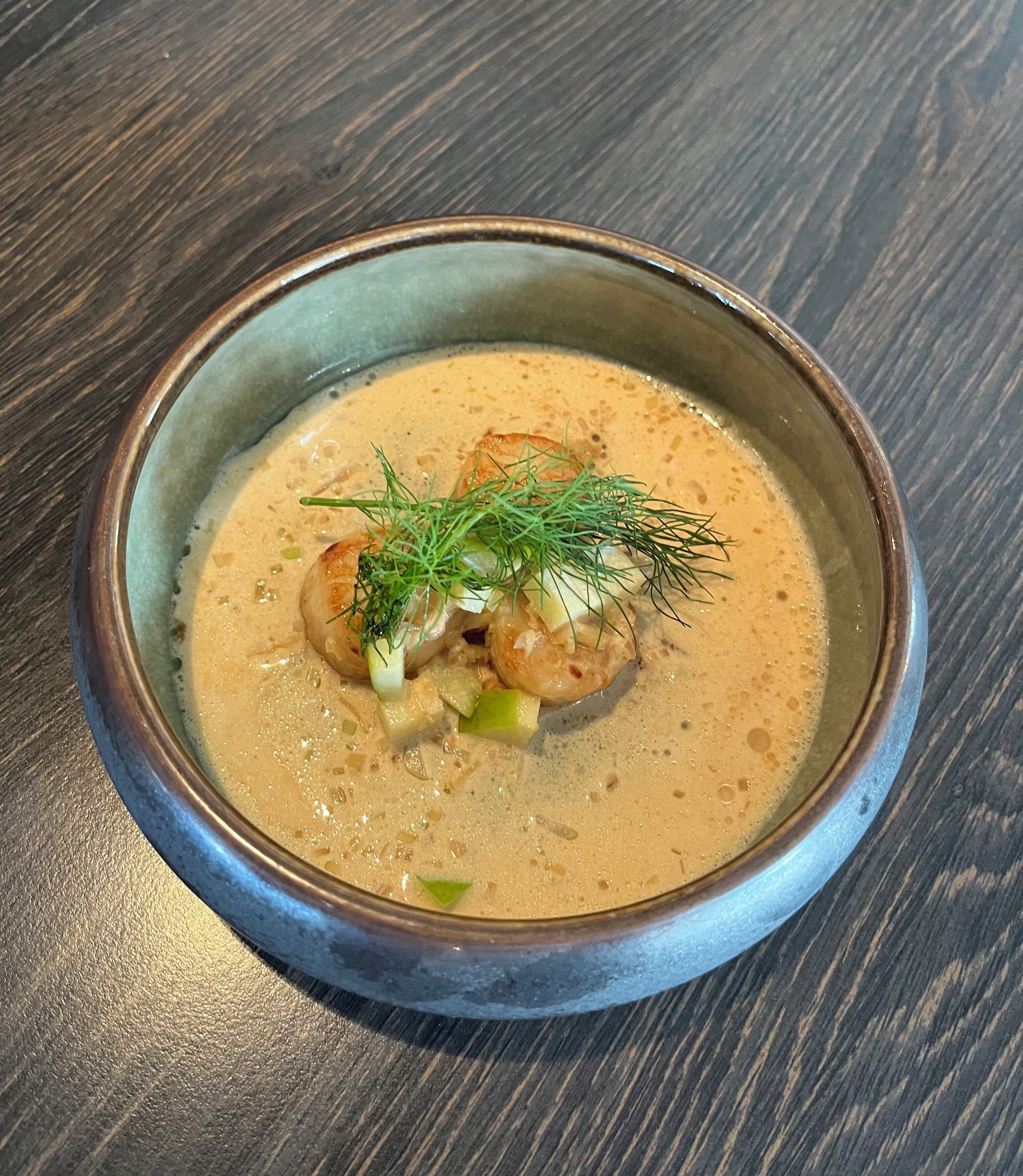 Un tazón de sopa con base cremosa, adornado con eneldo y vegetales picados, servido en un tazón de cerámica sobre una mesa de madera.
